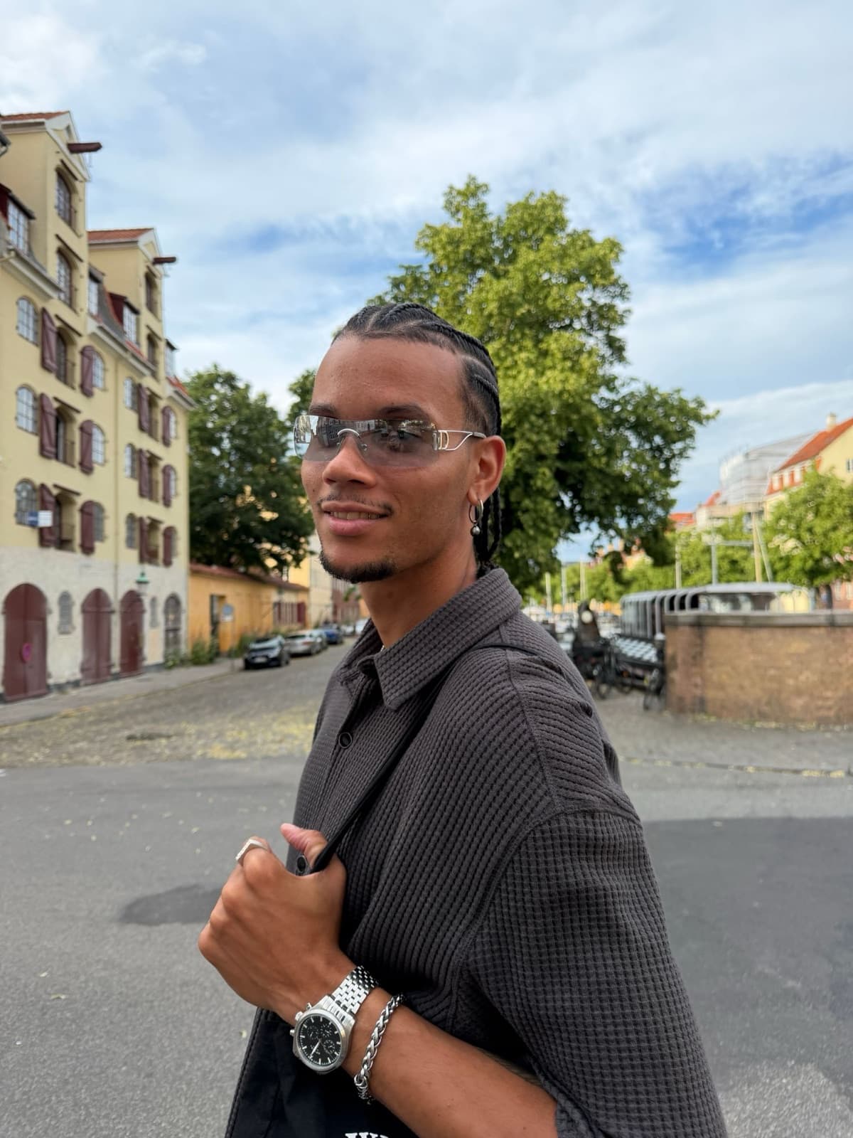 Remy smiling and looking back over one shoulder on a sunny street, wearing sunglasses and a dark shirt